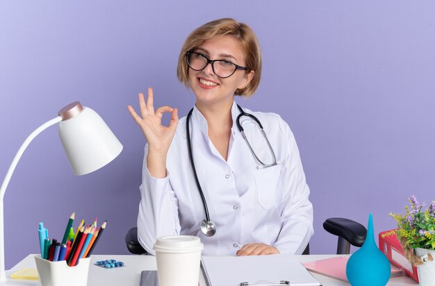 Lächelnde junge Ärztin, die ein medizinisches Gewand mit Stethoskop und Brille trägt, sitzt am Tisch mit medizinischen Instrumenten, die eine gute Geste einzeln auf blauem Hintergrund zeigen