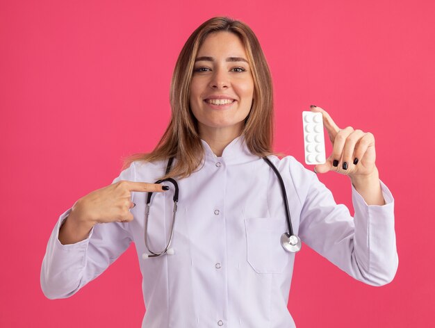 Lächelnde junge Ärztin, die medizinische Robe mit Stethoskop hält und Punkte auf Pillen isoliert auf rosa Wand trägt
