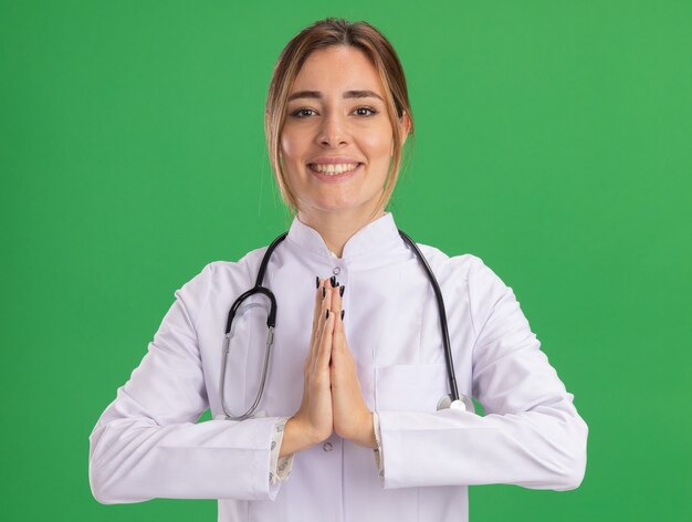 Lächelnde junge Ärztin, die medizinische Robe mit Stethoskop trägt, die Gebetsgeste lokalisiert auf grüner Wand zeigt