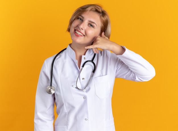 Lächelnde junge Ärztin, die medizinische Robe mit Stethoskop trägt, die Telefonanrufgeste einzeln auf orangefarbenem Hintergrund zeigt