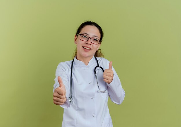 Lächelnde junge Ärztin, die medizinische Robe und Stethoskop mit Brille trägt, die Daumen oben isoliert zeigt
