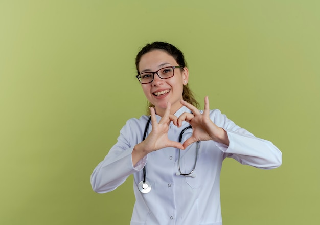 Lächelnde junge Ärztin, die medizinische Robe und Stethoskop mit Brille trägt, die Herzgeste lokalisiert auf olivgrüner Wand zeigt