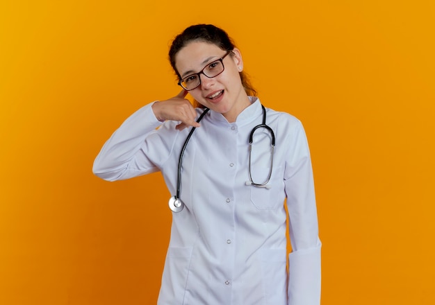 Lächelnde junge Ärztin, die medizinische Robe und Stethoskop mit Brille trägt, die Telefonanrufgeste lokalisiert auf orange Wand zeigt