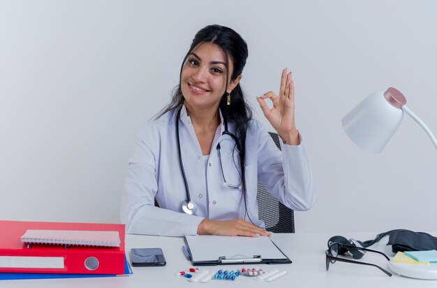 Lächelnde junge Ärztin, die medizinische Robe und Stethoskop trägt, sitzt am Schreibtisch mit medizinischen Werkzeugen, die die Hand auf den Schreibtisch setzen, die ok Zeichen lokalisiert tut