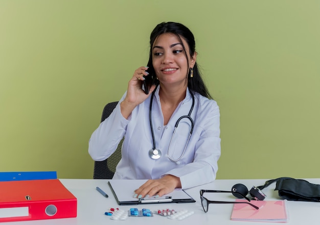 Lächelnde junge Ärztin, die medizinische Robe und Stethoskop trägt, sitzt am Schreibtisch mit medizinischen Werkzeugen, die Seite betrachten Hand auf Schreibtisch sprechen am Telefon lokalisiert betrachten