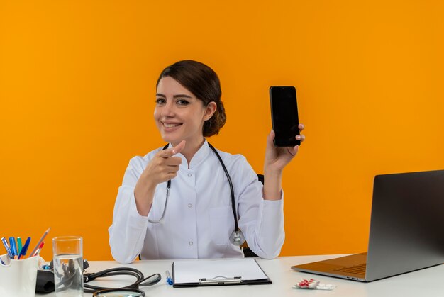 Lächelnde junge Ärztin, die medizinische Robe und Stethoskop trägt, sitzt am Schreibtisch mit medizinischen Werkzeugen und Laptop, der Handy zeigt, das lokal auf gelbe Wand zeigt