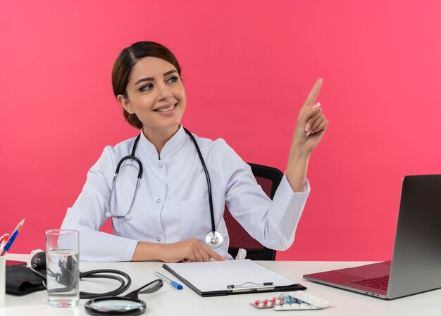Lächelnde junge Ärztin, die medizinische Robe und Stethoskop trägt, sitzt am Schreibtisch mit medizinischen Werkzeugen und Laptop, die auf Seite lokalisiert auf rosa Wand suchen und zeigen