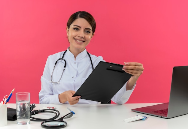 Lächelnde junge Ärztin, die medizinische Robe und Stethoskop trägt, sitzt am Schreibtisch mit medizinischen Werkzeugen und Laptop, die Klemmbrett lokalisiert auf rosa Wand halten