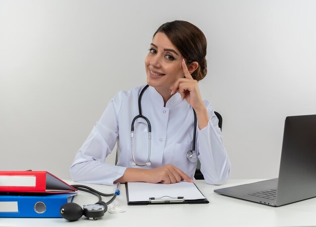 Lächelnde junge Ärztin, die medizinische Robe und Stethoskop trägt, sitzt am Schreibtisch mit medizinischen Werkzeugen und Laptop, die Kopf mit Finger lokalisiert auf weißer Wand berühren