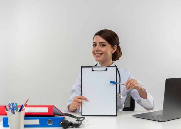 Lächelnde junge Ärztin, die medizinische Robe und Stethoskop trägt, sitzt am Schreibtisch mit medizinischen Werkzeugen und Laptop, die Zwischenablage halten, die mit Stift lokalisiert zeigt