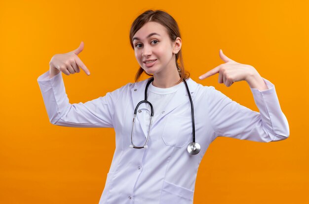 Lächelnde junge Ärztin, die medizinische Robe und Stethoskop trägt, zeigt auf sich selbst auf isolierter orange Wand