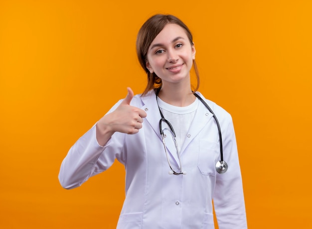 Lächelnde junge Ärztin, die medizinische Robe und Stethoskop trägt, zeigt Daumen oben auf isolierter orange Wand