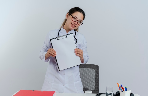 Lächelnde junge Ärztin, die medizinische Robe und Stethoskop und Brille trägt, die hinter Schreibtisch mit medizinischen Werkzeugen stehen, die die Zwischenablage lokalisiert suchen