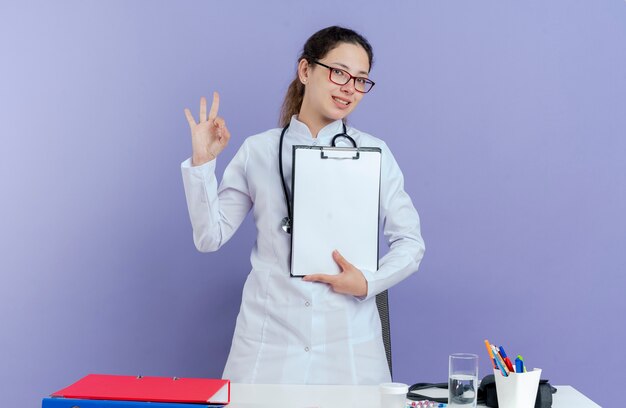 Lächelnde junge Ärztin, die medizinische Robe und Stethoskop und Brille trägt, die hinter Schreibtisch mit medizinischen Werkzeugen stehen, die Klemmbrett halten, das ok Zeichen tut, das isoliert schaut