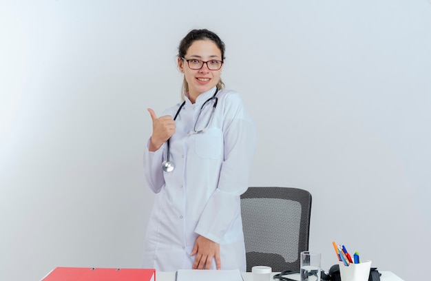 Lächelnde junge Ärztin, die medizinische Robe und Stethoskop und Brille trägt, die hinter Schreibtisch mit medizinischen Werkzeugen stehen, die zeigen, Daumen oben lokalisiert