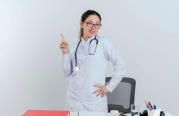 Lächelnde junge Ärztin, die medizinische Robe und Stethoskop und Brille trägt, die hinter Schreibtisch mit medizinischen Werkzeugen stehen und Hand auf Taille heben Finger lokalisiert suchen