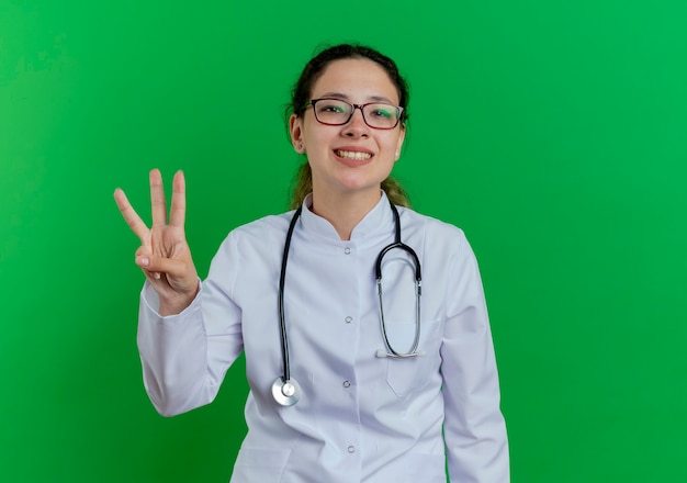 Lächelnde junge Ärztin, die medizinische Robe und Stethoskop und Brille trägt, zeigt drei mit der Hand lokalisiert auf grüner Wand mit Kopienraum