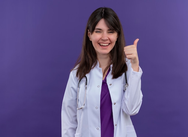 Lächelnde junge Ärztin im medizinischen Gewand mit Stethoskop-Daumen hoch auf lokalisiertem violettem Hintergrund mit Kopienraum