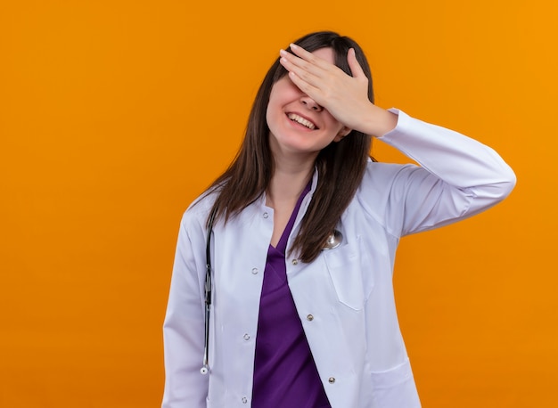 Lächelnde junge Ärztin im medizinischen Gewand mit Stethoskop legt Hand auf Augen auf lokalisiertem orange Hintergrund mit Kopienraum