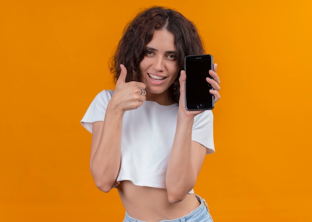 Lächelnde junge schöne Frau, die Handy hält und Daumen oben auf isolierter orange Wand mit Kopienraum zeigt