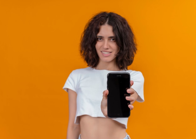 Lächelnde junge schöne Frau, die Handy hält und es auf isolierter orange Wand mit Kopienraum ausdehnt