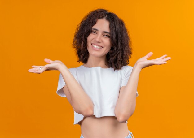 Lächelnde junge schöne Frau, die leere Hände auf isolierter orange Wand zeigt