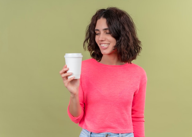 Lächelnde junge schöne Frau, die Plastikkaffeetasse hält und es auf isolierter grüner Wand mit Kopienraum betrachtet