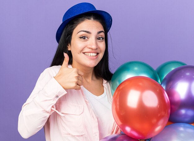 Lächelnde junge schöne Frau mit Partyhut, die Luftballons hält und Daumen nach oben zeigt, isoliert auf blauer Wand