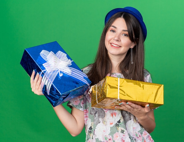 Lächelnde junge schöne Frau mit Partyhut mit Geschenkboxen isoliert auf grüner Wand