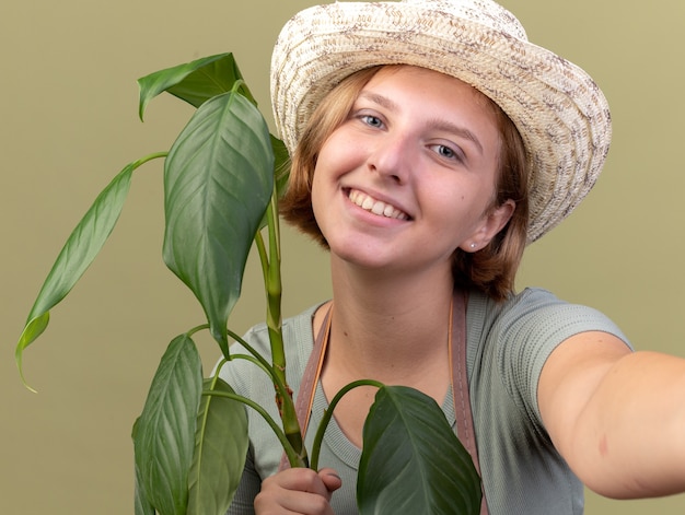 Lächelnde junge slawische Gärtnerin mit Gartenhut, die Pflanze hält und vorgibt, die Kamera zu halten, die ein Selfie macht