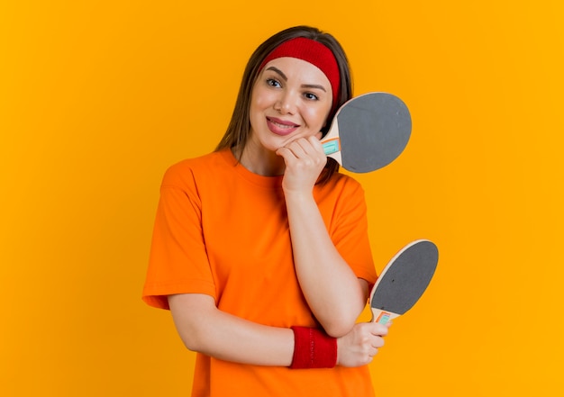 Lächelnde junge sportliche Frau, die Stirnband und Armbänder hält, die Tischtennisschläger halten, die Hand auf Kinn setzen und Seite betrachten