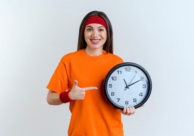 Lächelnde junge sportliche Frau, die Stirnband und Armbänder hält und auf Uhr lokalisiert auf weißer Wand mit Kopienraum trägt