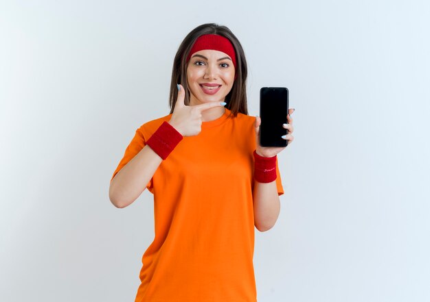 Lächelnde junge sportliche Frau, die Stirnband und Armbänder trägt, zeigt Handy, das auf sie schaut, die lokal suchen