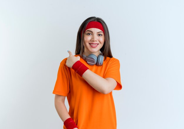 Lächelnde junge sportliche Frau, die Stirnband und Armbänder und Kopfhörer am Hals trägt und hinter isoliert zeigt