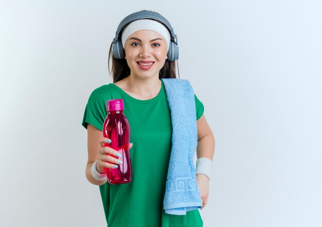 Lächelnde junge sportliche Frau, die Stirnband und Armbänder und Kopfhörer hält, die Wasserflasche mit Handtuch auf Schulter lokalisiert auf weißer Wand mit Kopienraum halten