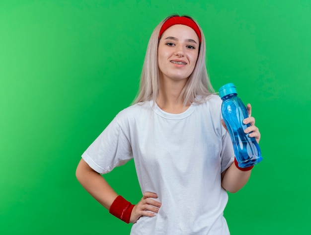 Lächelnde junge sportliche Frau mit Zahnspangen, die Stirnband und Armbänder tragen, hält Wasserflasche und legt Hand auf Taille lokalisiert auf grüner Wand