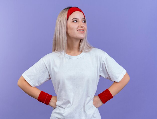 Lächelnde junge sportliche Frau mit Zahnspangen, die Stirnband und Armbänder tragen, legt Hände auf Taille und betrachtet Seite isoliert auf lila Wand