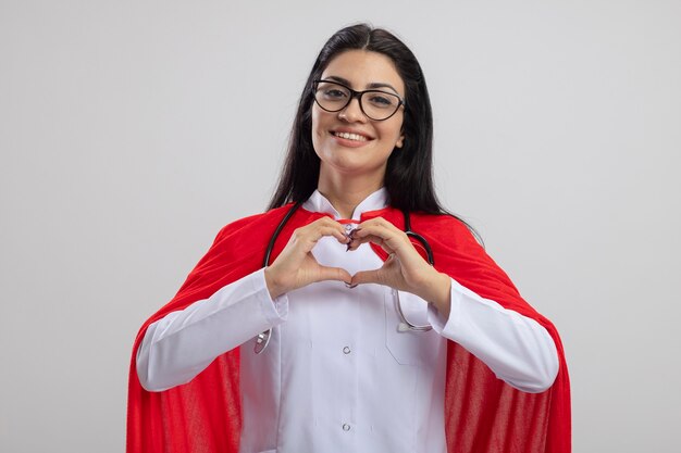 Lächelnde junge Superfrau, die Brille und Stethoskop trägt, die Front tut, die Herzzeichen lokalisiert auf weißer Wand tut