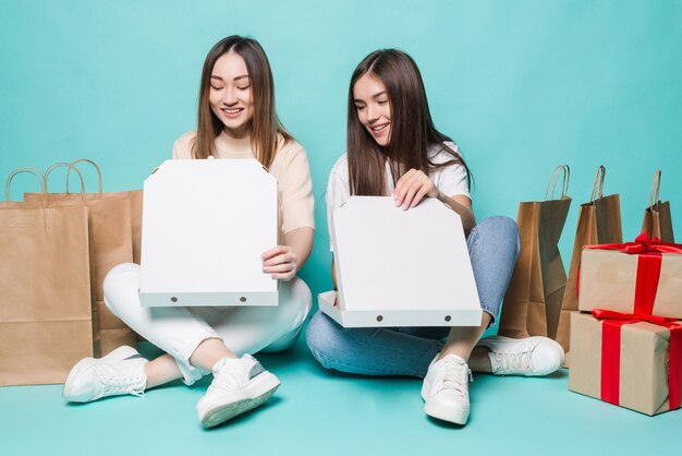 Lächelnde junge zwei Mädchen sitzen Boden Einkaufstüten und Geschenk offene Pizza auf der türkisfarbenen Wand.