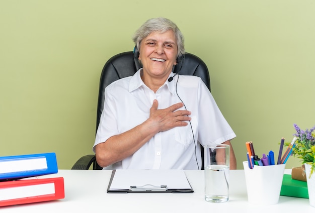 Lächelnde kaukasische Callcenter-Betreiberin auf Kopfhörern, die am Schreibtisch mit Bürowerkzeugen sitzen und die Hand auf ihre Brust legen, isoliert auf grüner Wand