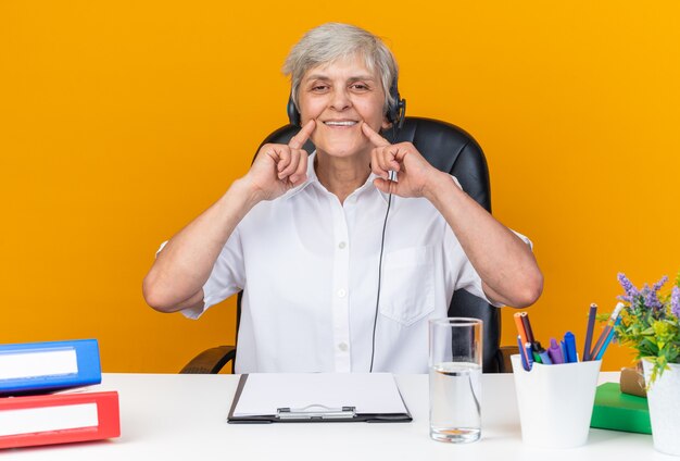 Lächelnde kaukasische Callcenter-Betreiberin auf Kopfhörern, die am Schreibtisch mit Bürowerkzeugen sitzen und ihr Lächeln mit den Fingern halten, die auf orangefarbener Wand isoliert sind