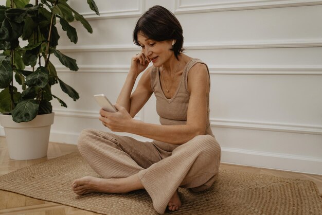 Lächelnde kaukasische erwachsene brünette Frau, die sich zu Hause mit Handy und Kopfhörern auf dem Teppich ausruht Konzept der positiven Emotionen
