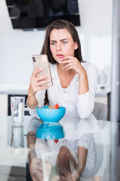 Lächelnde kaukasische Frau, die Handy verwendet, während frisches Gemüsesalat in der Küche essen