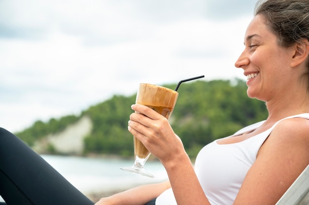 Lächelnde kaukasische Frau, die Kaffeegetränk an einem Strand mit Schaum und Trinkhalm mit Hügeln hält