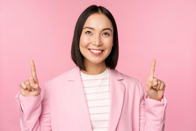 Lächelnde koreanische Geschäftsfrau, die mit dem Finger nach oben zeigt und Werbebanner oder Logo auf der Oberseite zeigt, die im Anzug über rosa Hintergrund steht