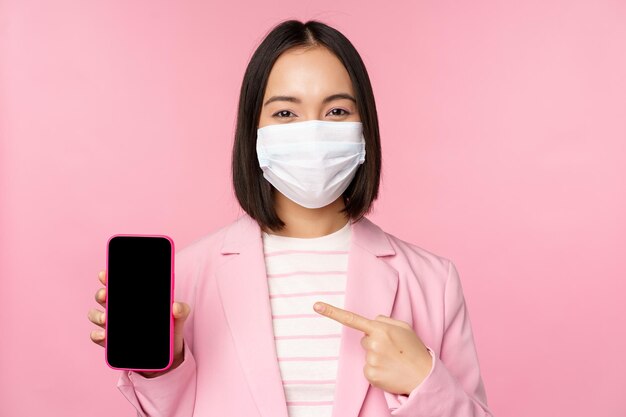 Lächelnde koreanische Geschäftsfrau im Anzug mit medizinischer Gesichtsmaske von Covid, die mit dem Finger auf den Handybildschirm zeigt, der über rosa Hintergrund steht