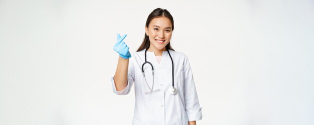 Lächelnde koreanische Ärztin zeigt Fingerherzgeste in Gummihandschuhen, die eine medizinische Uniform tragen und glücklich auf den weißen Hintergrund der Kamera schauen