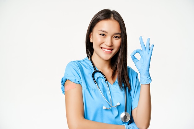 Lächelnde Krankenschwester, asiatische Ärztin in Peelings, zeigt ein OK-Zeichen und einen geimpften Arm mit medizinischem Pflaster, empfiehlt die Impfung von Covid-19, weißer Hintergrund