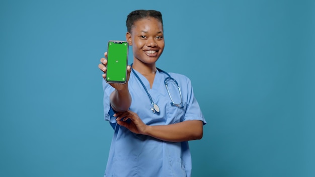 Lächelnde krankenschwester, die grünen bildschirm auf dem handy an der kamera zeigt. arzthelferin in uniform mit smartphone mit chroma-key und isolierter mockup-vorlage, stehend auf blauem hintergrund.