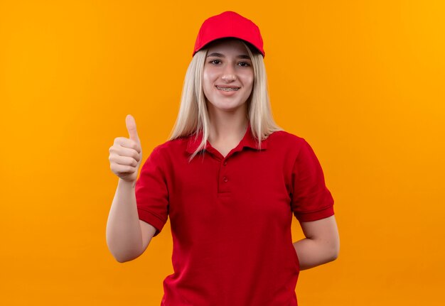 Lächelnde Lieferung junges Mädchen, das rotes T-Shirt und Kappe in Zahnspange ihren Daumen oben auf lokalisiertem orange Hintergrund trägt